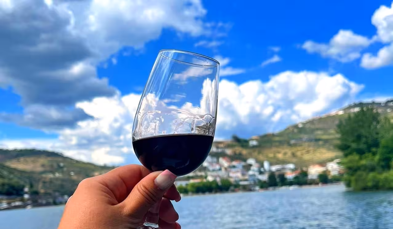 Prestige Tour wine tasting by the river, hand holding red wine glass with scenic hillside village view