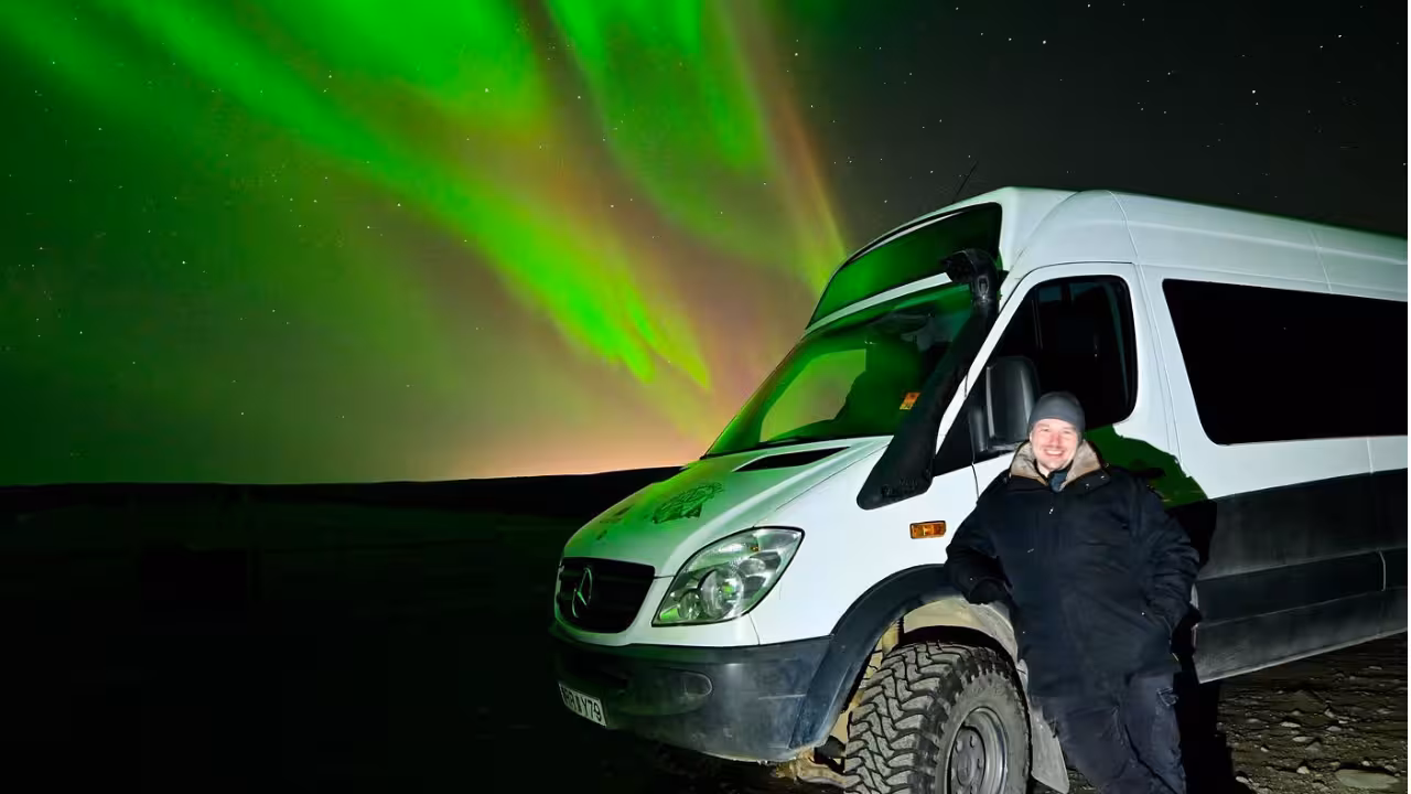 Smiling guide standing beside a 4x4 van under vivid green northern lights on a private Iceland aurora tour from Reykjavik