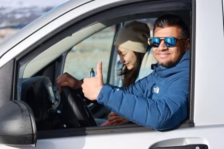 Smiling driver giving thumbs up from white transfer vehicle with passenger in back seat