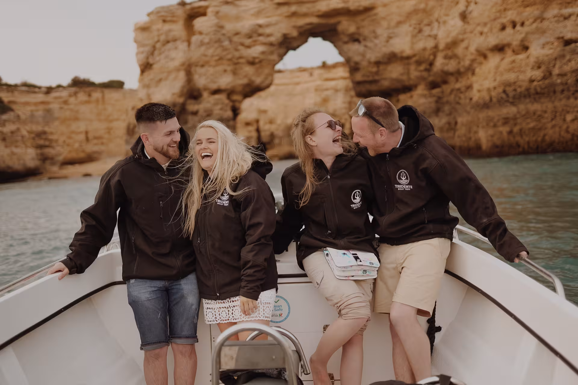 Friends laughing on a private Benagil Caves sunset boat tour, wearing Tridente jackets with Algarve sea cliffs behind