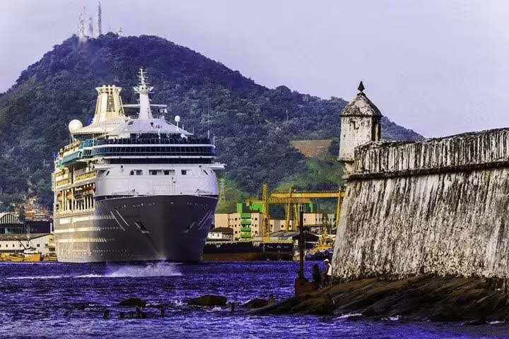 Cruise ship navigating past historic fortifications with a mountainous backdrop in Santos harbor.