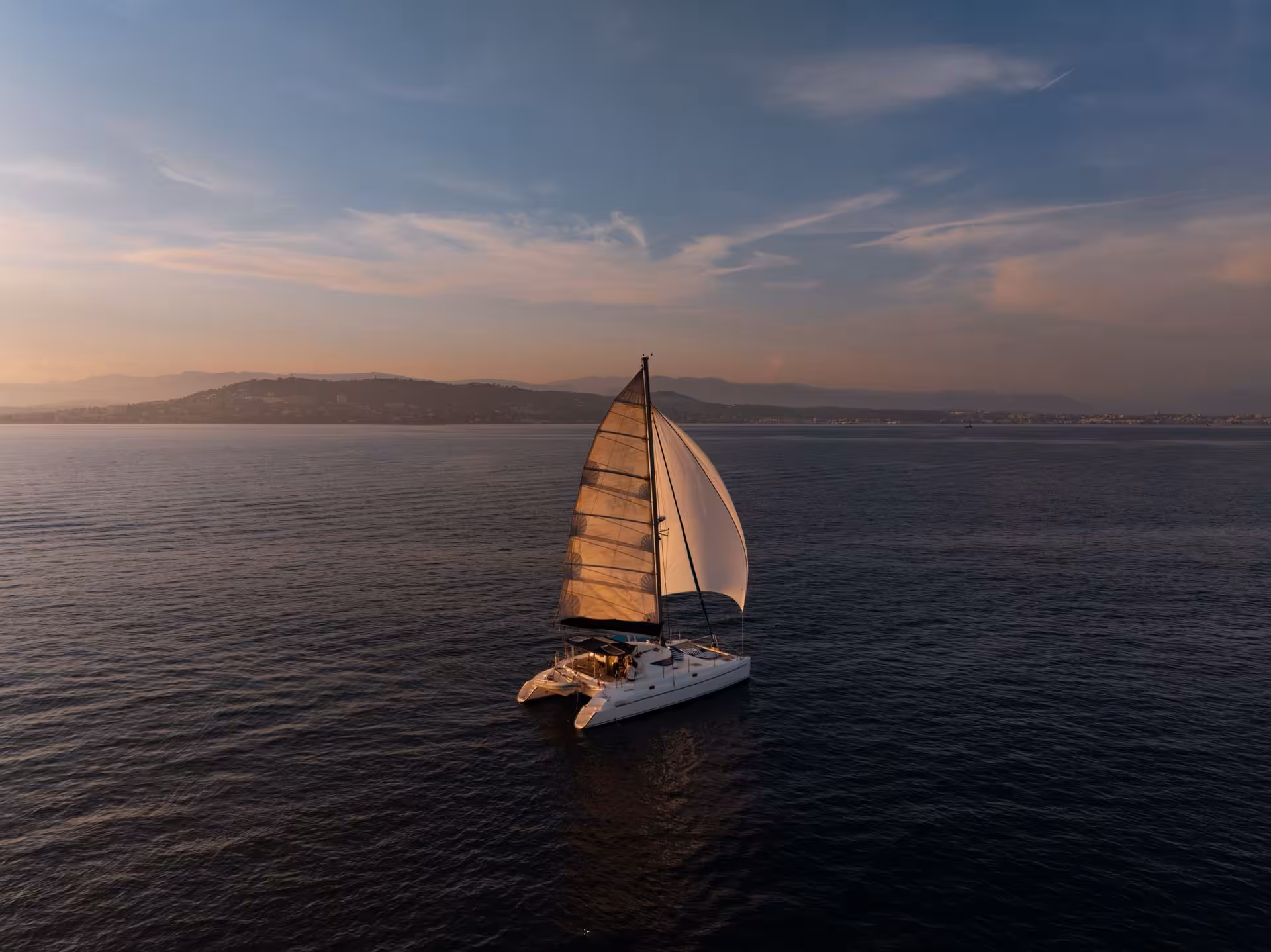 White sailing catamaran on calm sea at sunset near Cannes with golden sails and hilly coastline