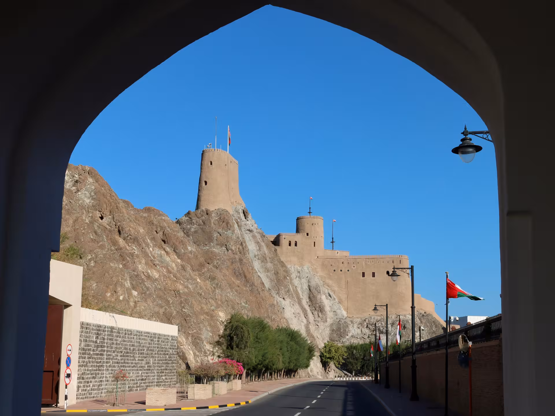 Private chauffeur service in Muscat driving past historic hilltop fort and Omani flags on a clear blue-sky day