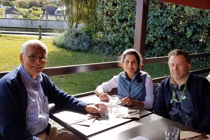 Three people smiling at outdoor restaurant table with green garden backdrop on Normandy day trip