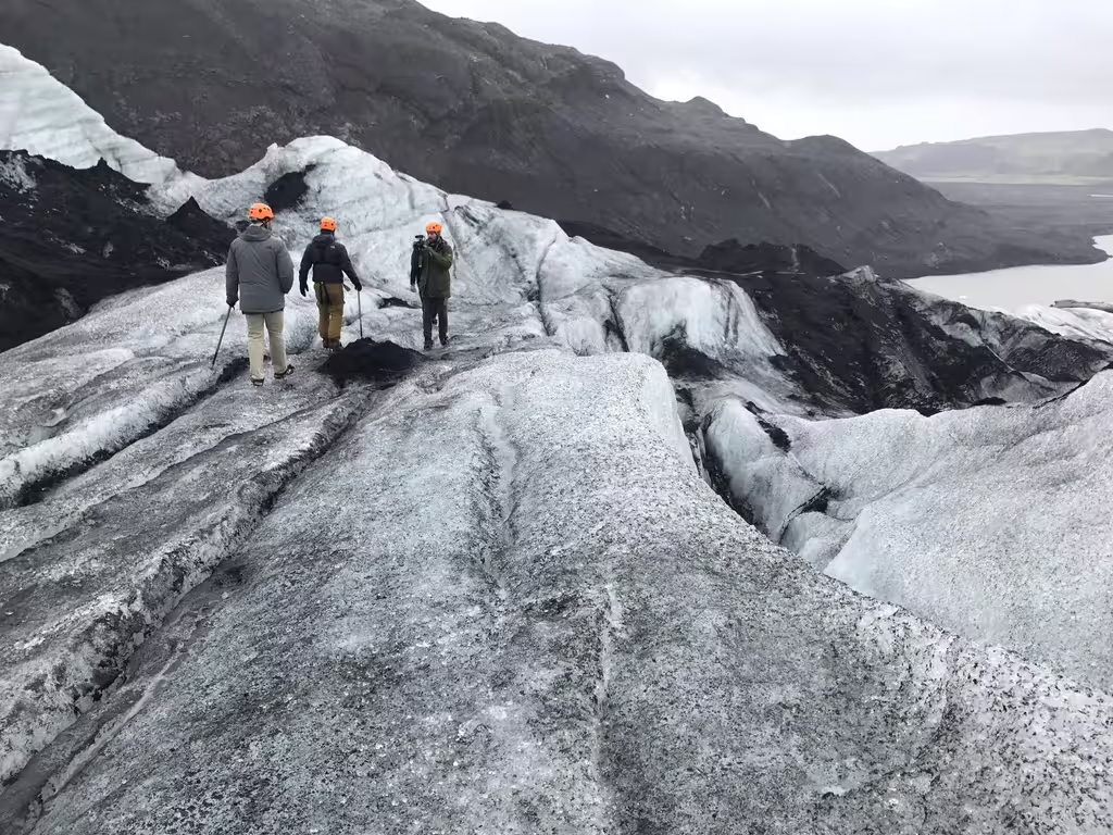 Three hikers in orange helmets walking on a glacier with black volcanic mountains and lake in background