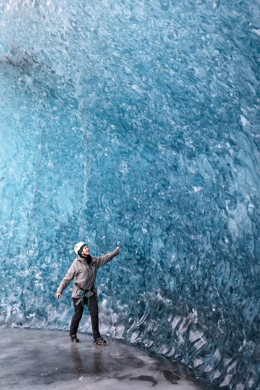 A smiling adventurer in a white helmet and harness points at the massive swirling blue ice cave wall during a private glacier photoshoot tour.