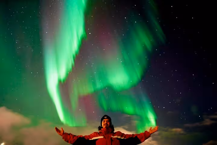 Person in red jacket with arms outstretched under vivid green northern lights in Reykjavik night sky