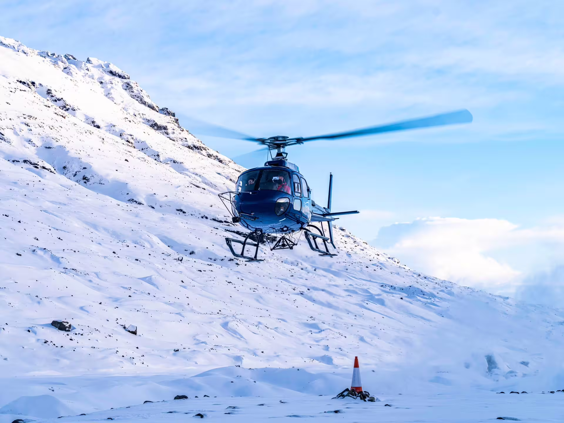 Blue helicopter hovering over snowy glacier slopes near Skaftafell with snow-capped mountains in background