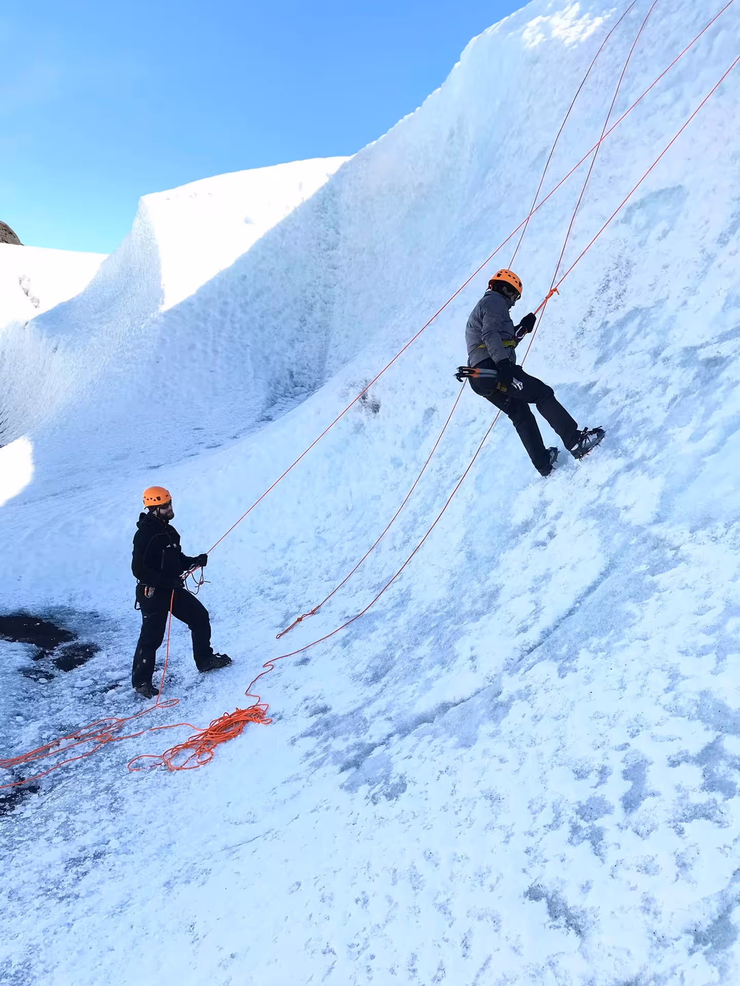 Two climbers in orange helmets ice climbing a steep snow wall on Solheimajokull Glacier with red ropes