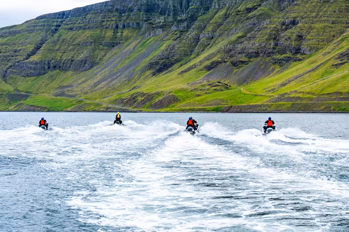 Four jet ski riders speeding across a fjord with green mountain cliffs in Iceland