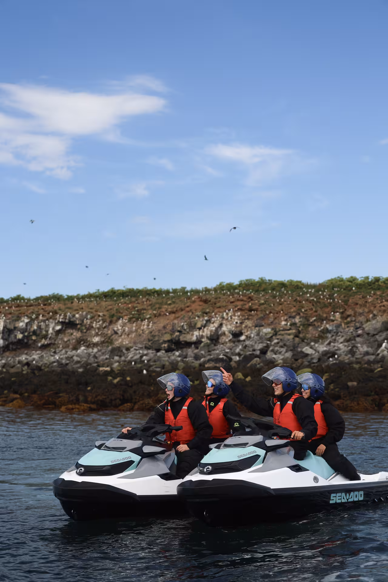 Four riders on Sea-Doo jet skis wearing helmets and life vests near a rocky island coastline