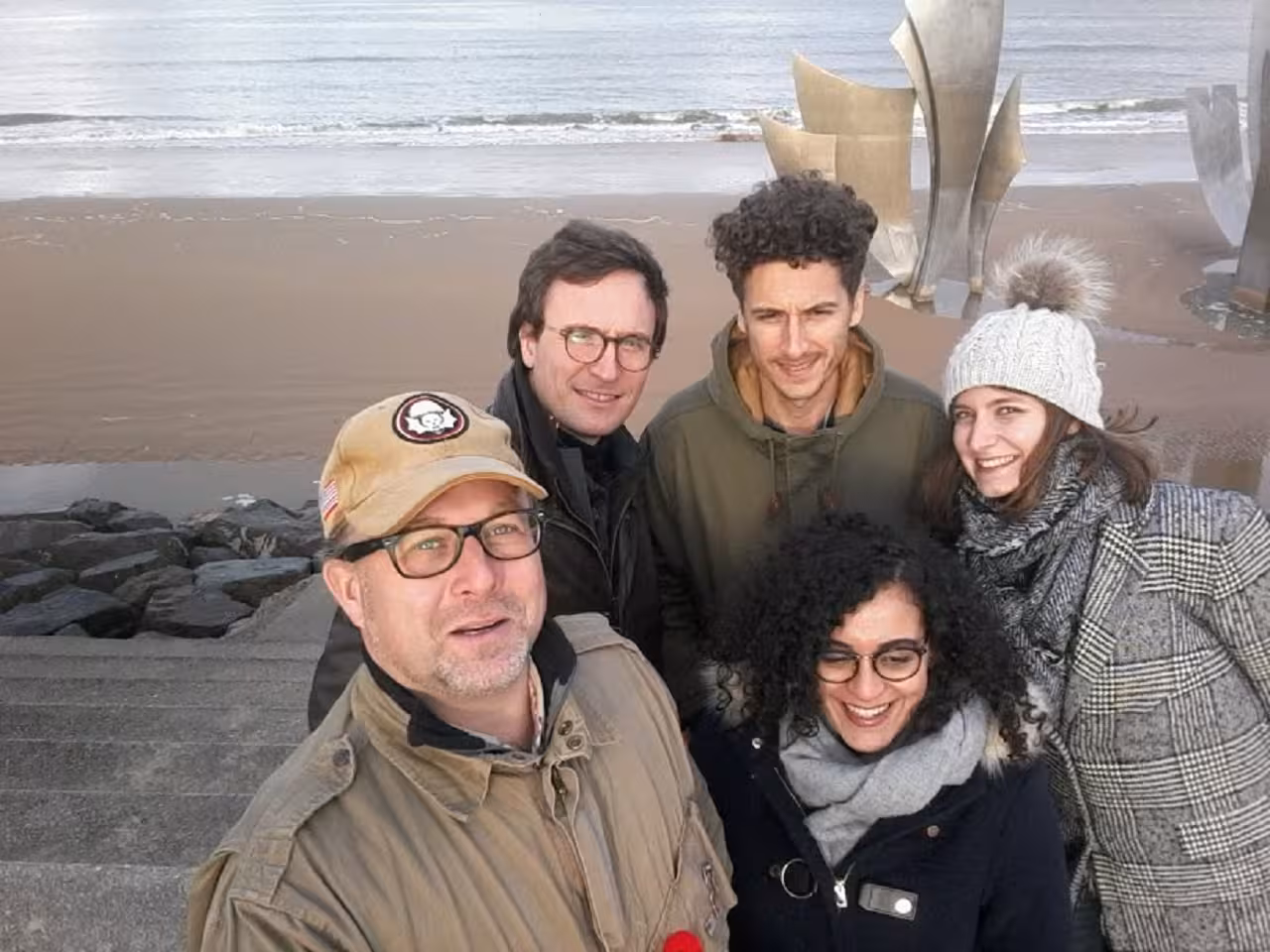 Group of five tourists posing for selfie on Normandy beach with metal sculpture and ocean in background