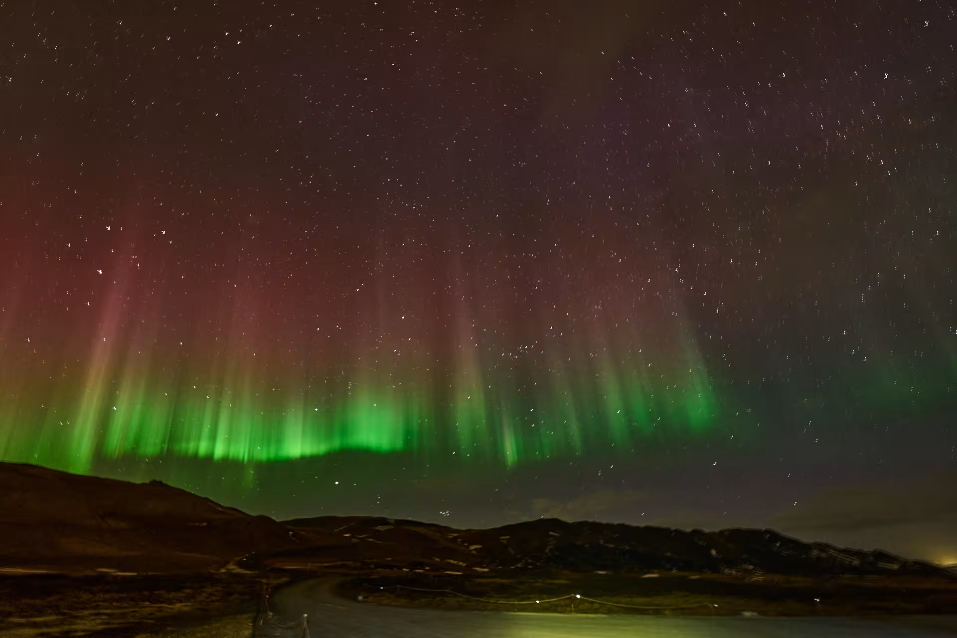 Vivid green and red aurora borealis curtains lighting up a starry night sky over dark hills