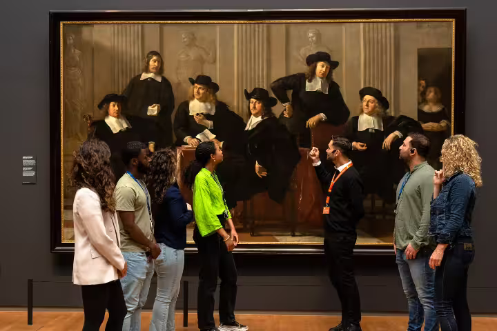 Visitors engage with a guide during a private tour at the Rijksmuseum, admiring a classic Dutch painting in Amsterdam.