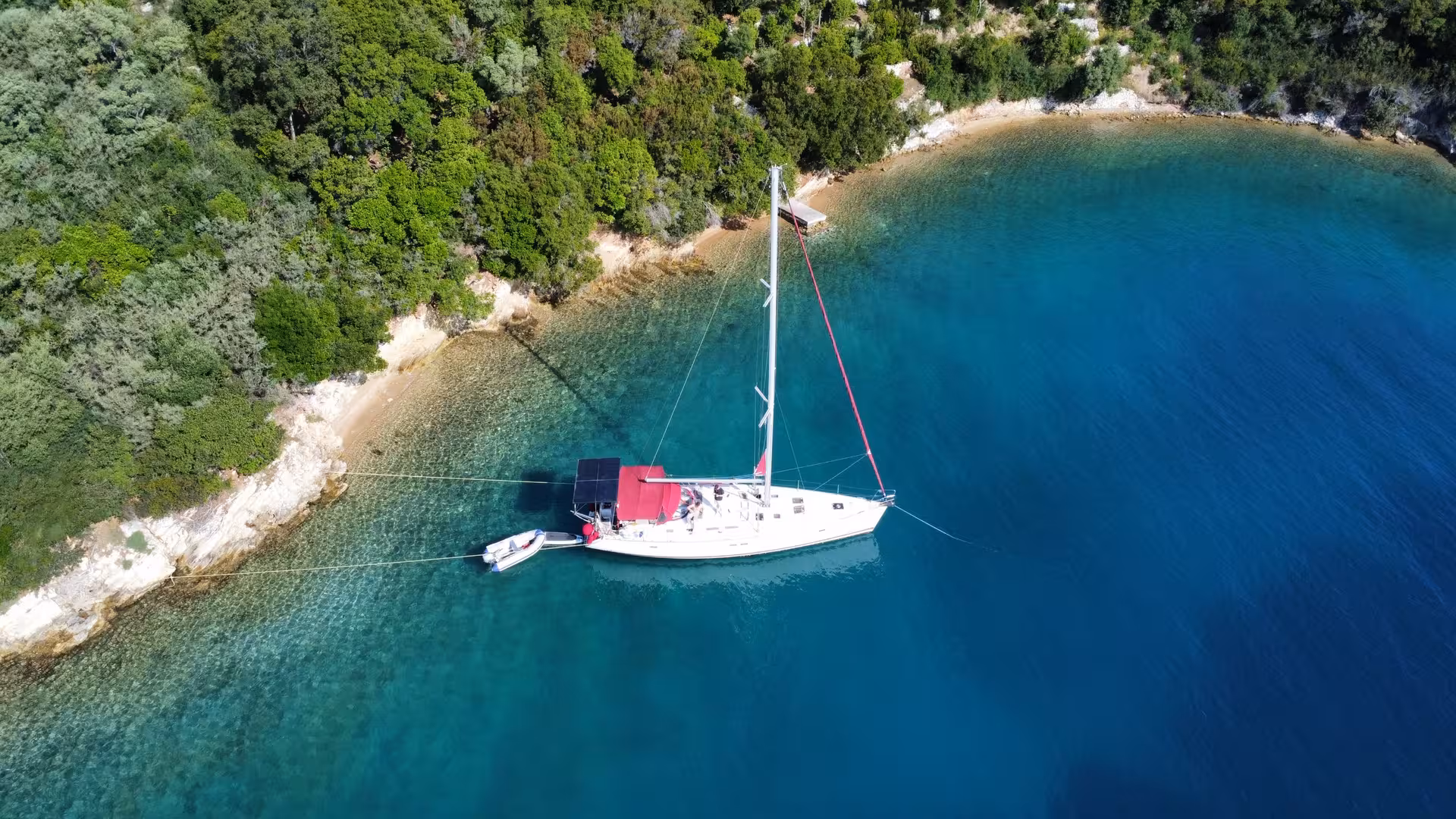 White sailboat anchored in turquoise bay near lush green coastline, aerial view
