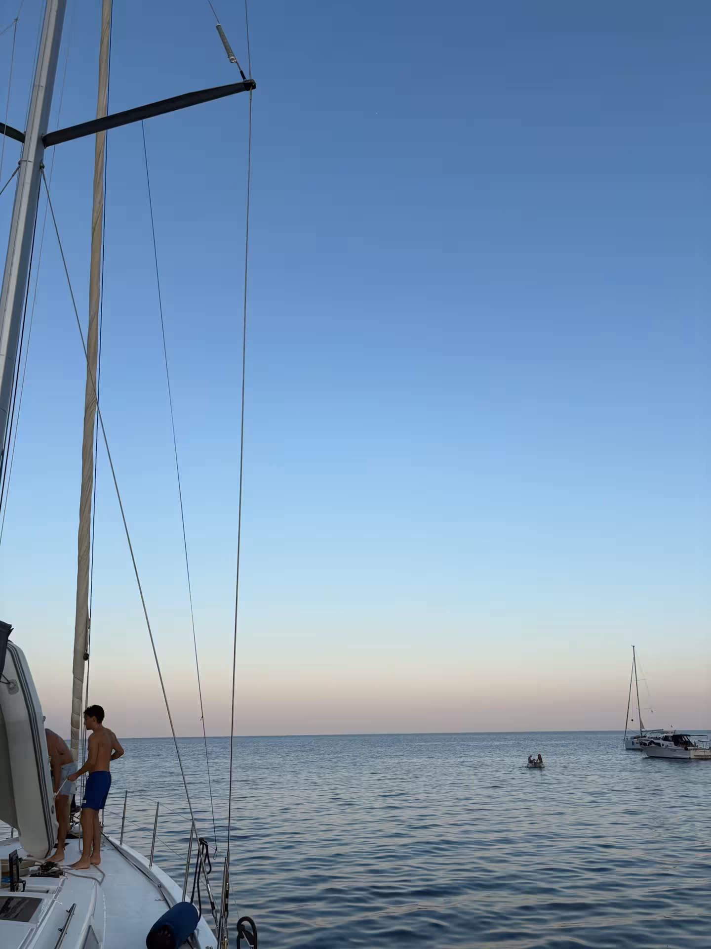 Shirtless sailor on deck of private sailboat at dusk with calm Ligurian Sea and anchored yacht in background