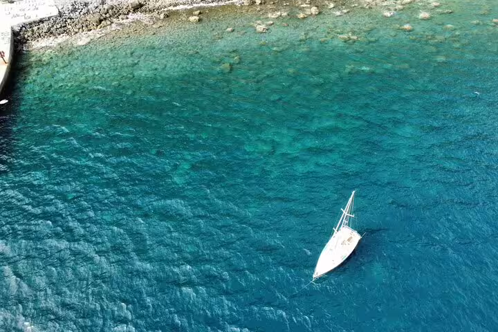 Aerial view of a private sailboat cruising on crystal-clear turquoise waters near a rocky shoreline, perfect for a full-day sailing adventure.