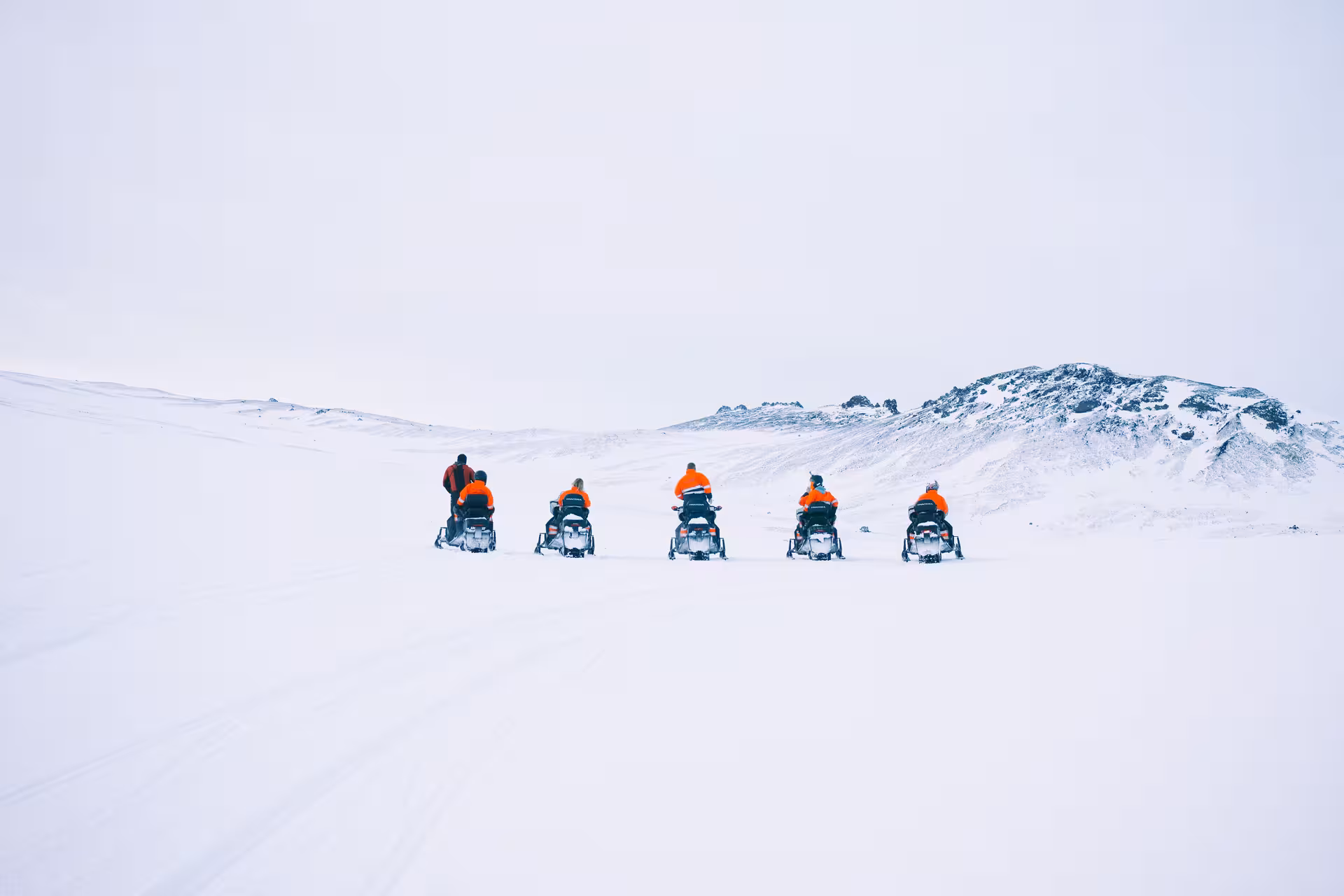 Group of snowmobilers in orange suits riding across snowy Eyjafjallajokull glacier toward rocky mountain