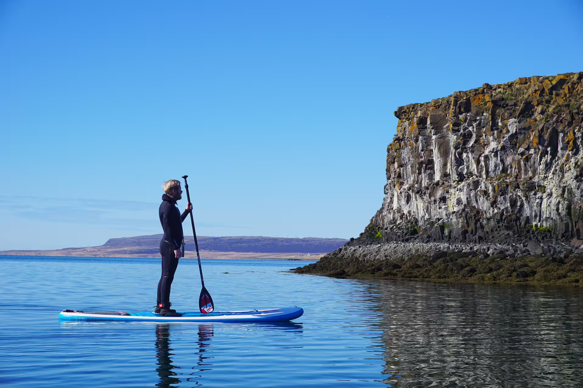 Paddle boarder explores serene waters beside rocky cliffs on a sunny day during a private SUP tour in the Forgotten Fjords.