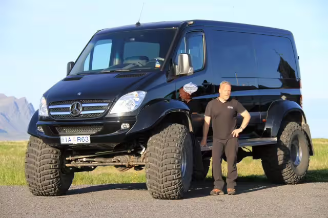 Man standing beside a black lifted Mercedes Sprinter Super Jeep on a paved road in Iceland
