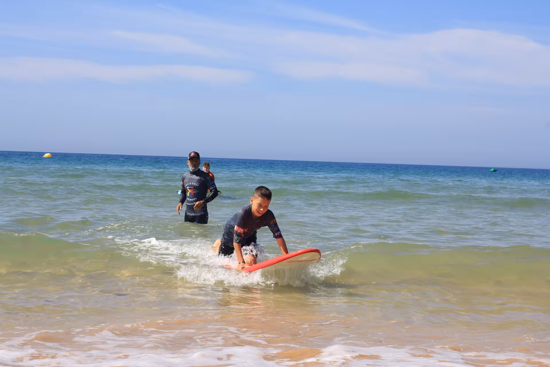 Surf instructor guiding a child on a soft board in shallow water during a safe, fun private surf lesson for kids on a sunny beach