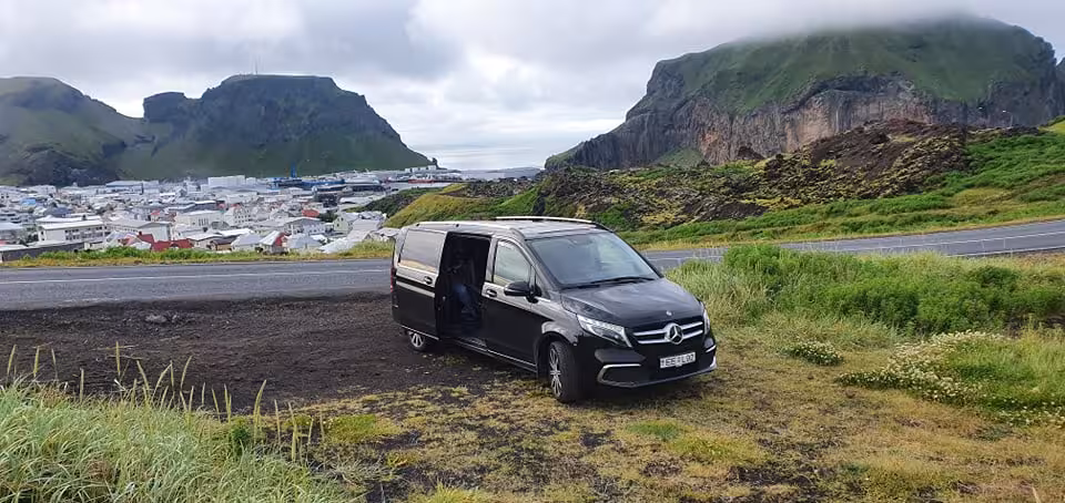Black Mercedes-Benz private van parked overlooking Westman Islands town with green volcanic mountains backdrop