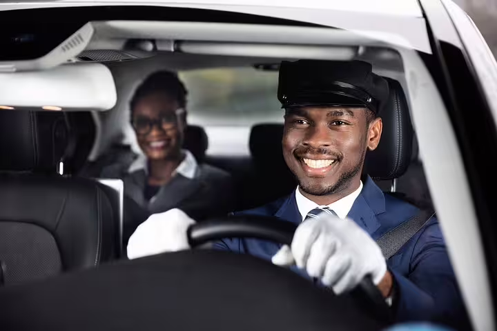 Smiling chauffeur in uniform and cap driving a private transfer car with passenger in back seat