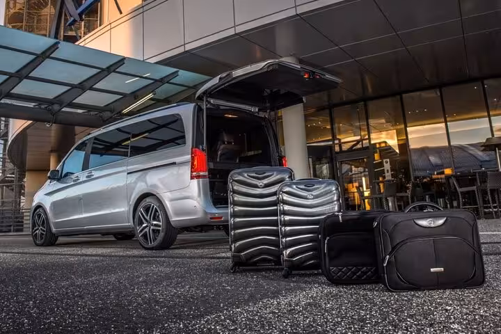 Silver Mercedes V-Class minivan with open trunk and black luggage outside modern hotel entrance