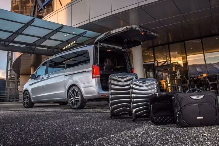 Silver Mercedes minivan with open trunk and black luggage outside modern airport terminal