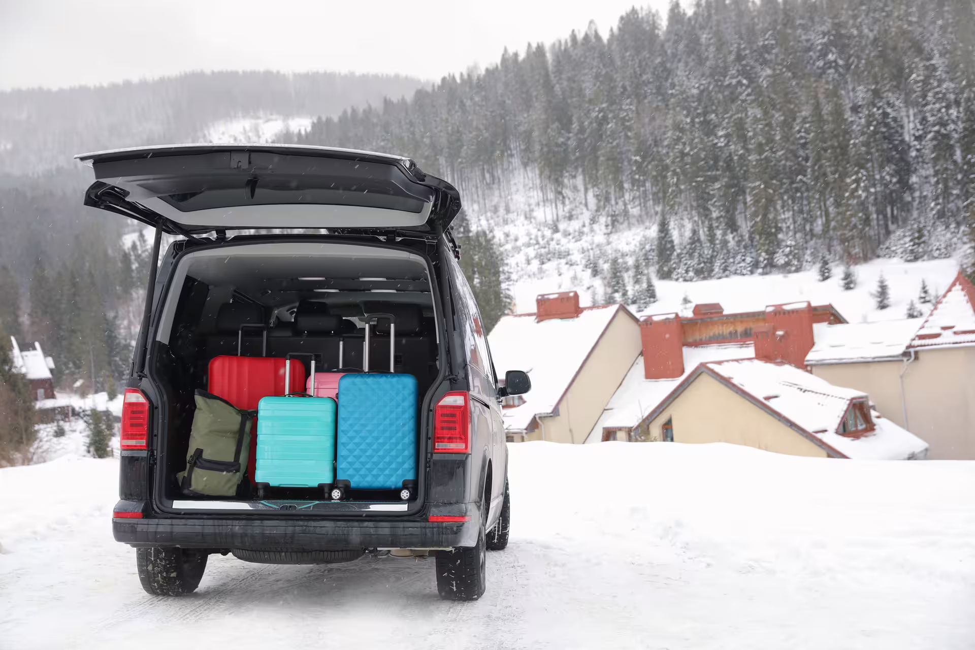 Black minivan with open trunk loaded with colorful suitcases on snowy mountain road in winter