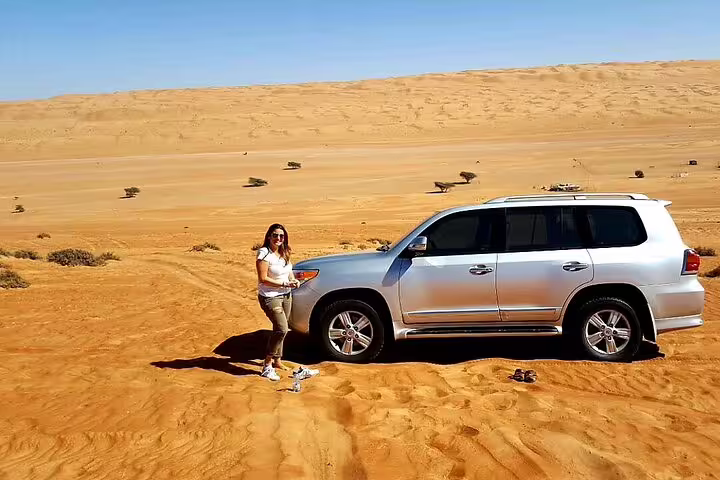 Woman enjoying the Wahiba Sands desert landscape beside a silver SUV, emphasizing private transfer from Muscat.
