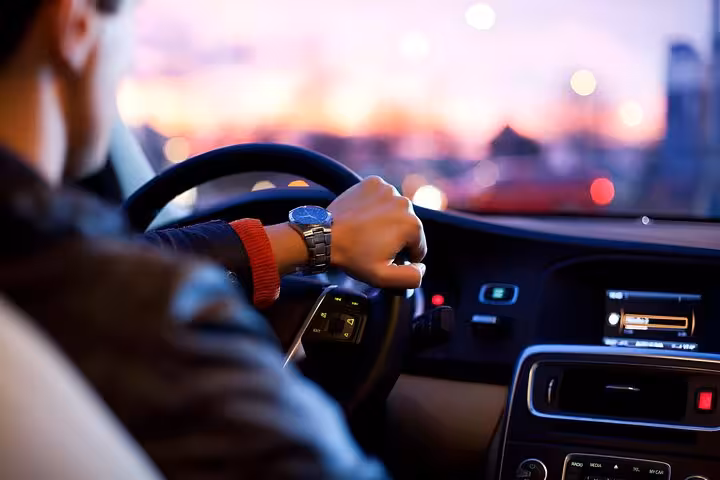 Driver wearing a blue watch steering a car at dusk with city lights in background