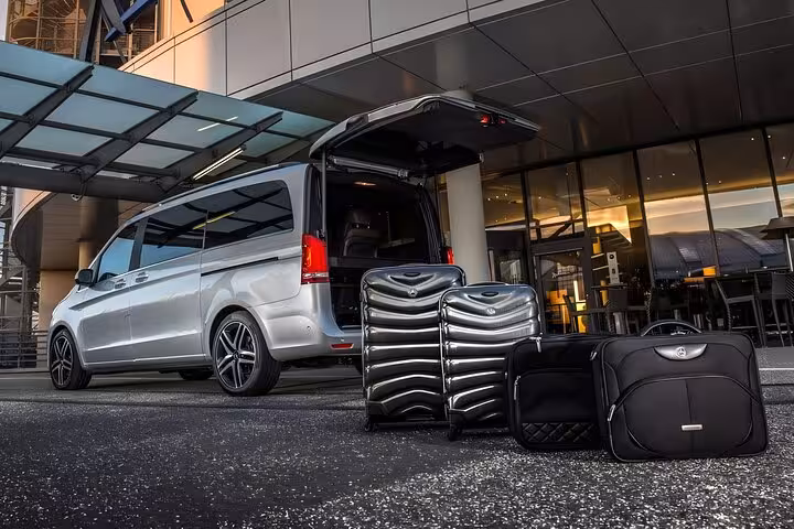 Silver Mercedes minivan with open trunk and black luggage outside modern hotel entrance