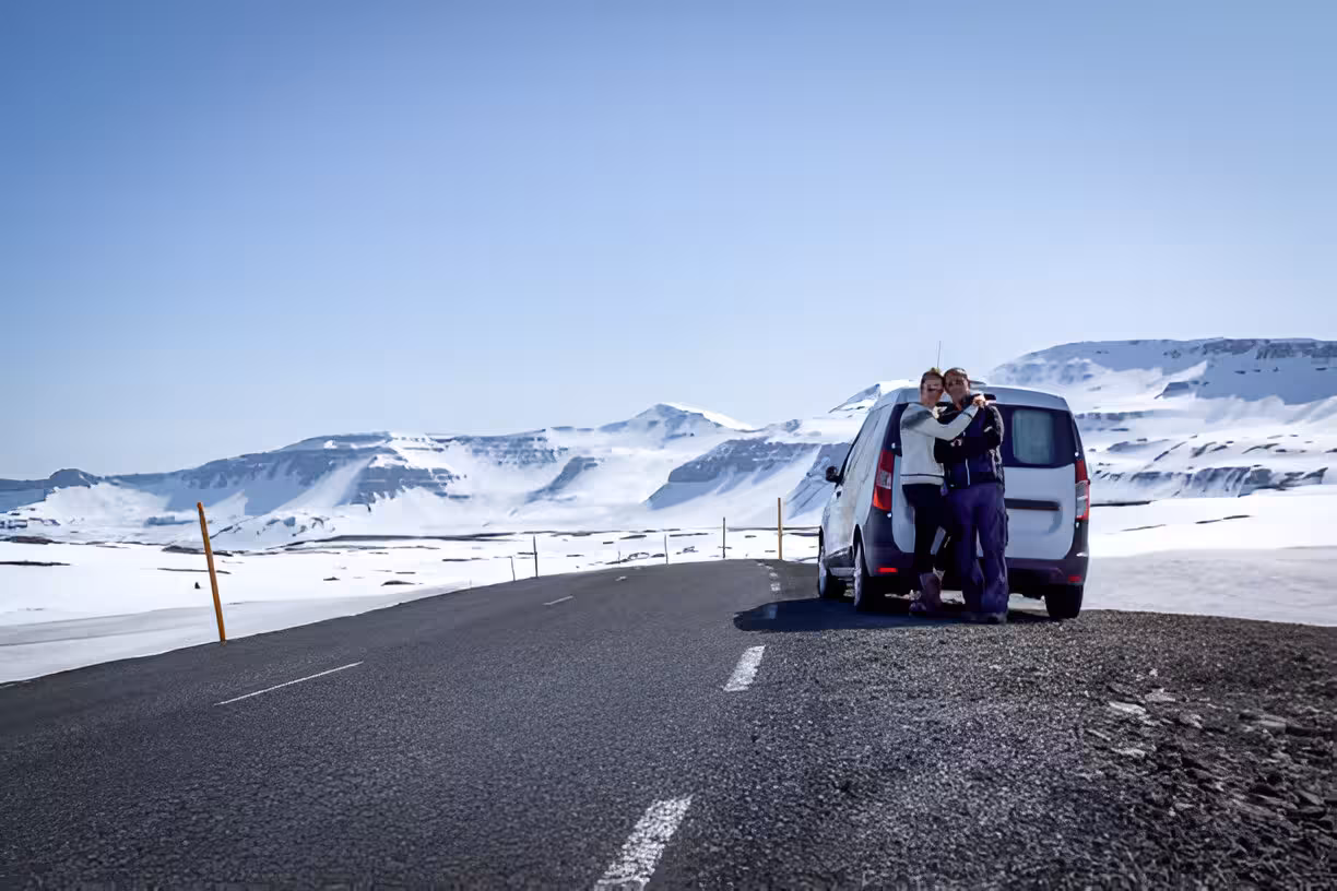 Two travelers posing beside a white van on a snowy Icelandic road with snow-covered mountains