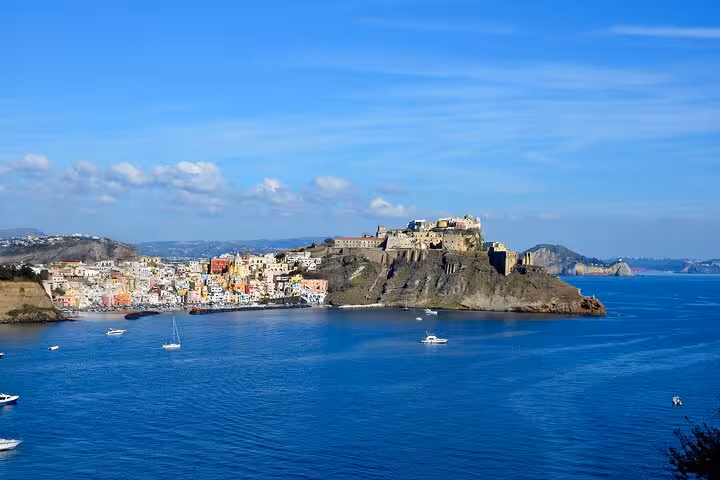 Stunning view of Procida's historic fortress and colorful town from the sea, with sailboats dotting the blue waters.