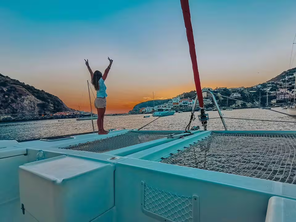 Sunset cruise to Procida by catamaran with guest celebrating on deck, overlooking colorful harbor villages and anchored yachts