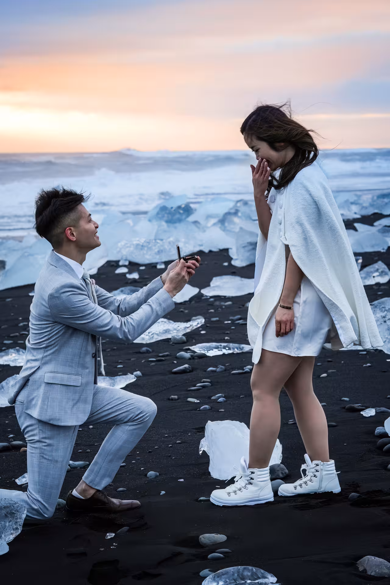 Man proposing on one knee at Iceland Diamond Beach with ice chunks and a breathtaking sunrise sky backdrop.
