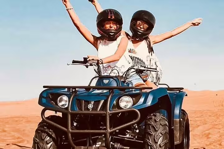 Two people enjoying an exhilarating quad bike ride on the red dunes during a desert safari adventure, wearing helmets and raising arms.
