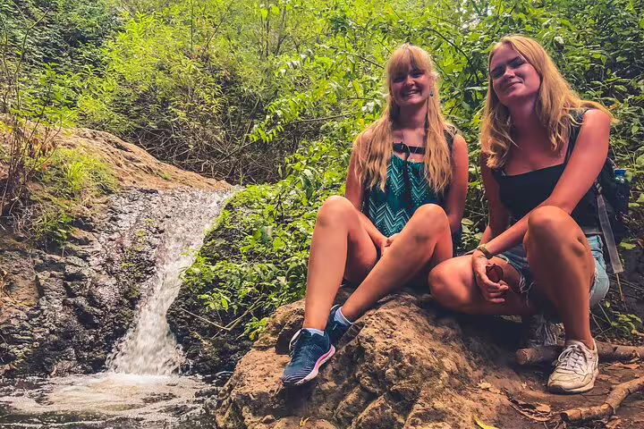 Two women sitting on rocks by a small waterfall in the rainforest, enjoying The Rainforest Tour experience.