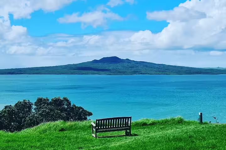 Scenic view of Rangitoto Island from Auckland with a bench on lush green grass under a vibrant blue sky.