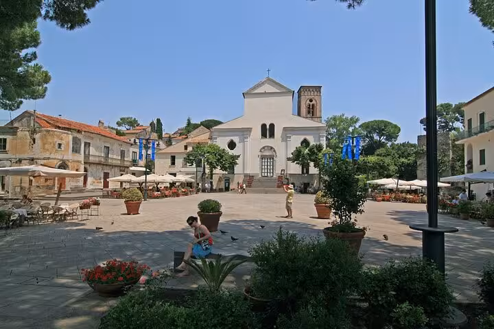 Charming Ravello town square with historic white church, surrounded by cafes and lush greenery under a clear sky.