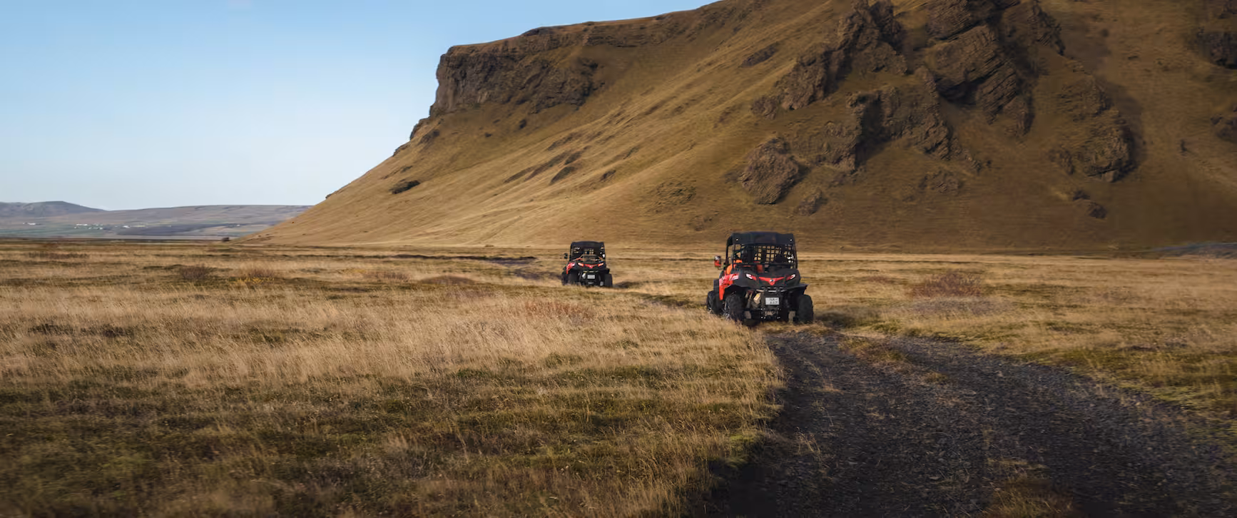 Two red off-road buggies driving on a dirt trail through golden grasslands with a dramatic Icelandic mountain backdrop