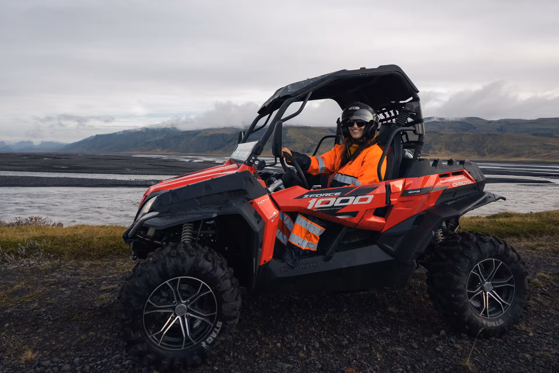 Smiling woman in orange safety gear driving red CFMOTO ZForce 1000 buggy on Iceland's volcanic landscape