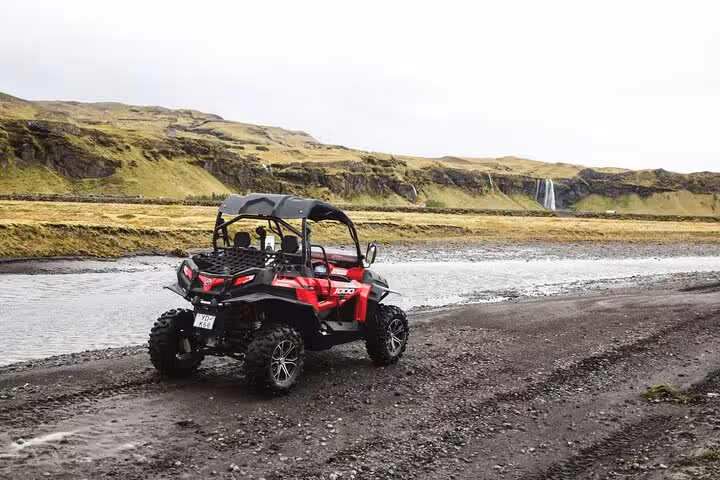 Red buggy UTV parked near a river with green Icelandic cliffs and a waterfall in the background