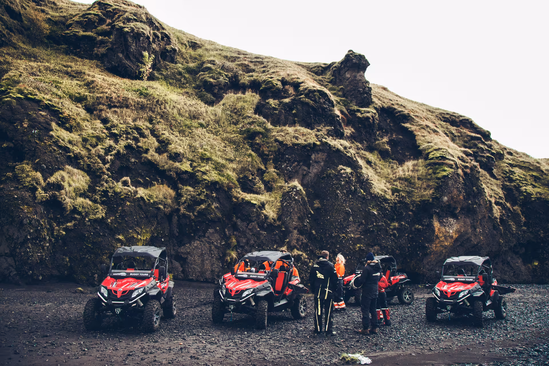 Red off-road buggy tour vehicles parked on Iceland's black sand beach below mossy volcanic cliffs