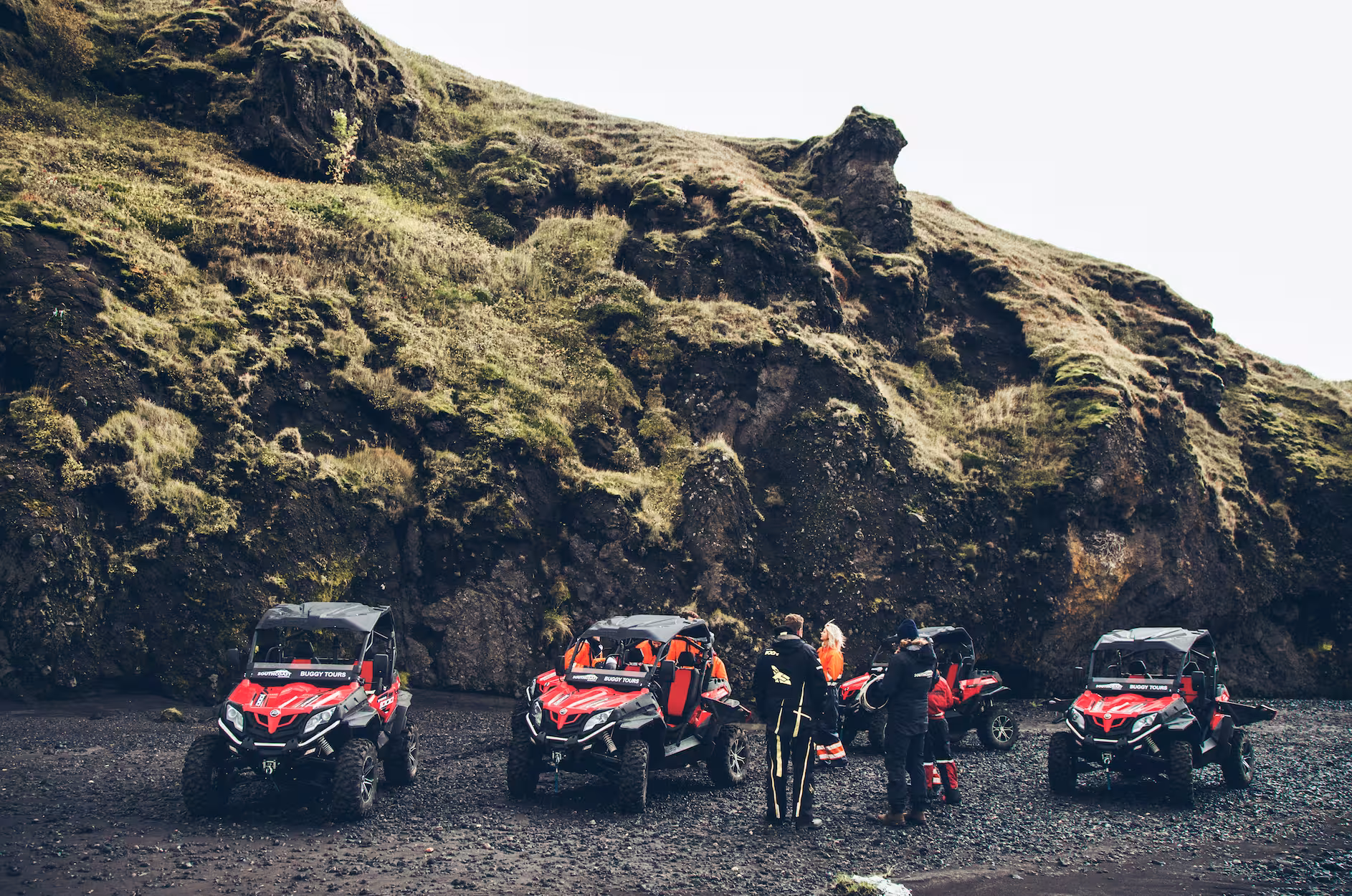 Group of tourists with red buggy tour vehicles parked on black sand beach below mossy volcanic cliffs in Iceland