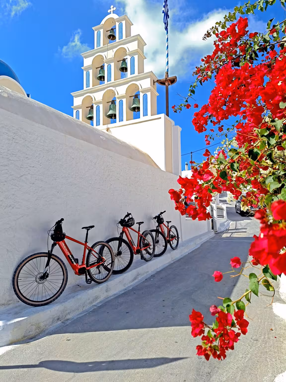 Red e-bikes on a Santorini e-bike tour by a white church bell tower and bougainvillea, scenic stop