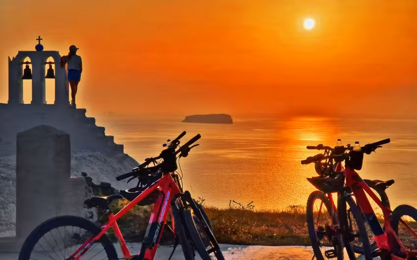 Red e-bikes by a Santorini chapel at sunset, overlooking the caldera on a guided e-bike sunset tour