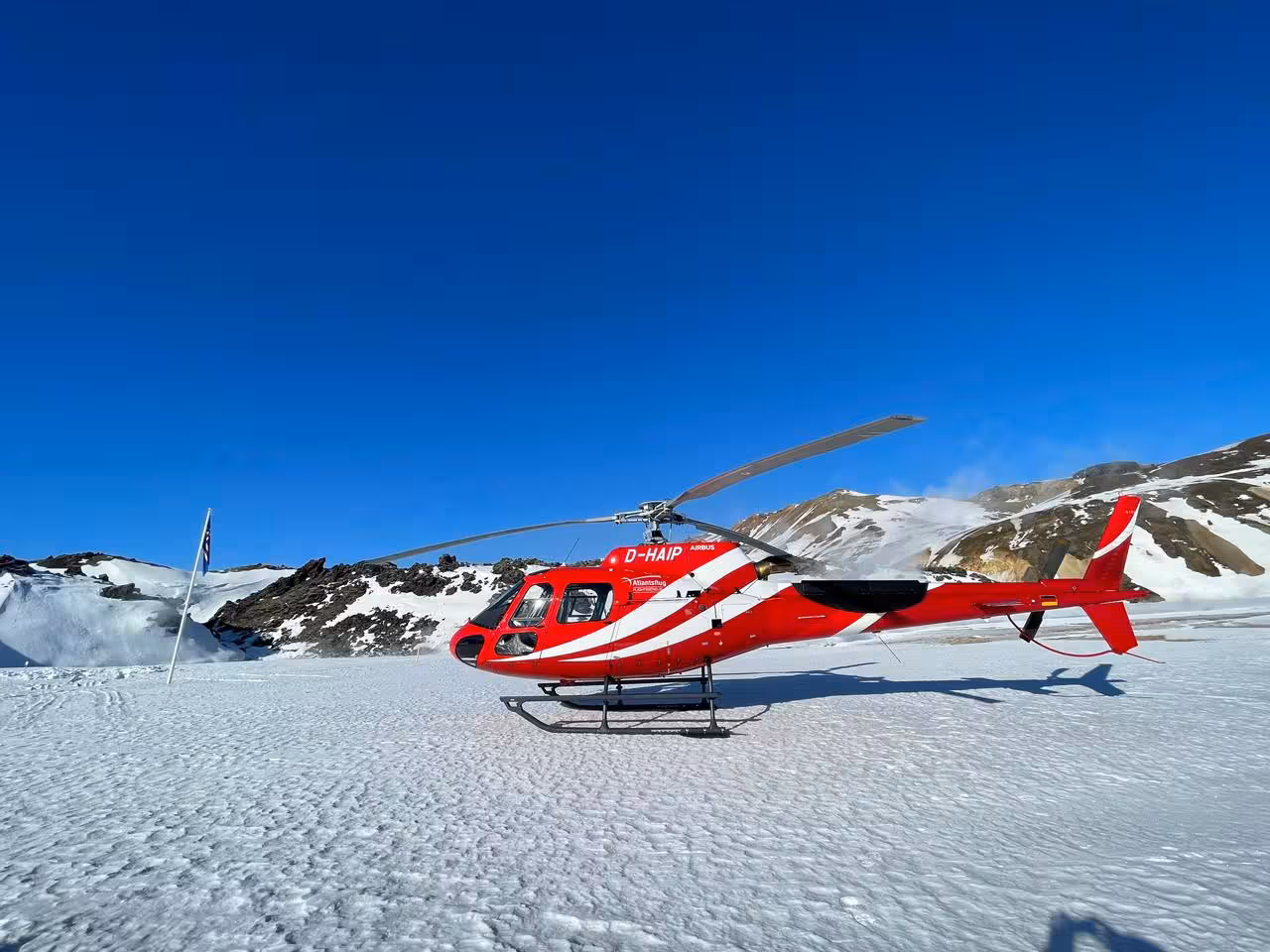 Red and white Airbus helicopter D-HAIP landed on a snow-covered glacier with snowy mountains behind