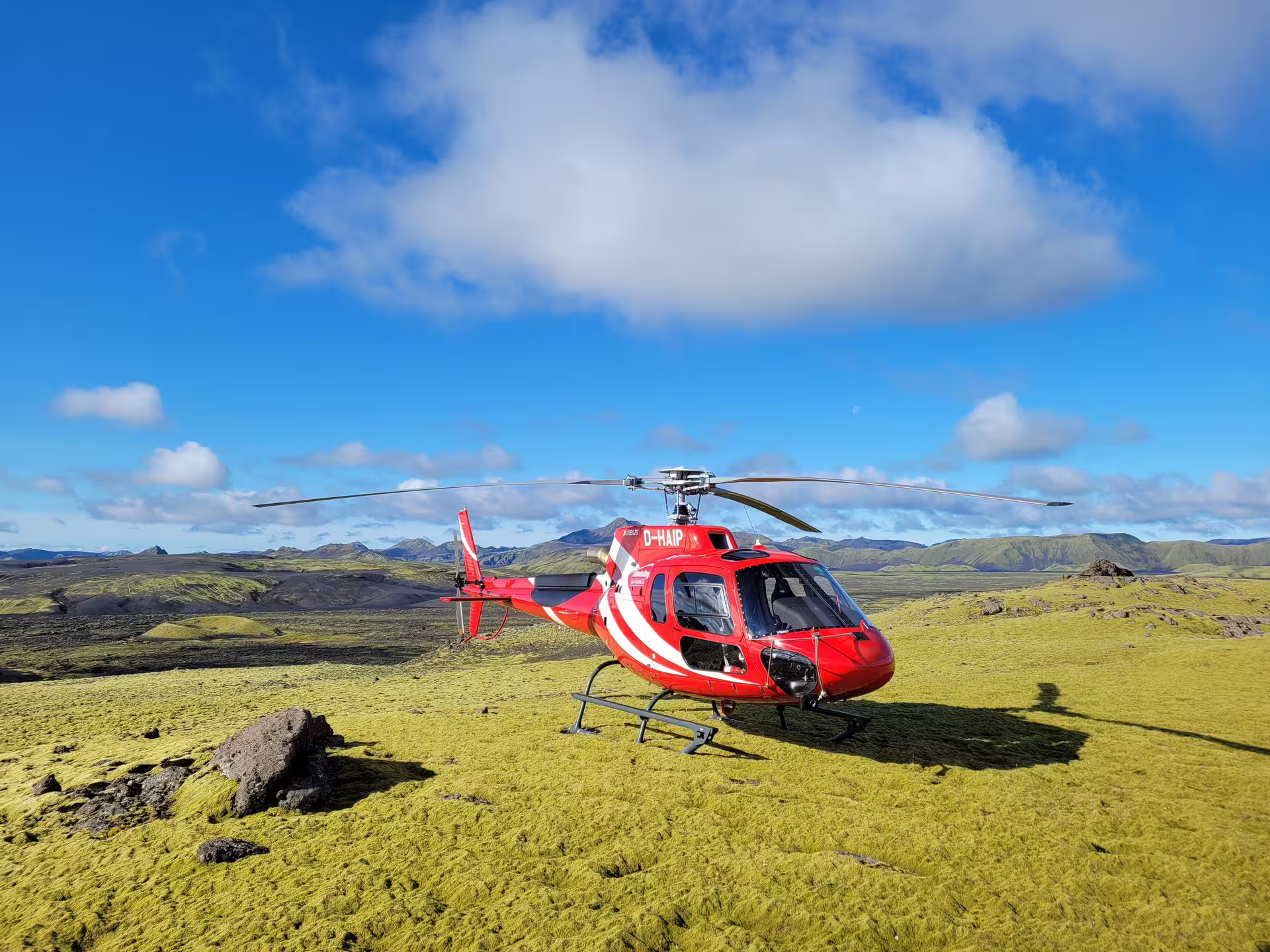 Red Airbus helicopter D-HAIP landed on green moss-covered lava fields with volcanic mountains in Iceland